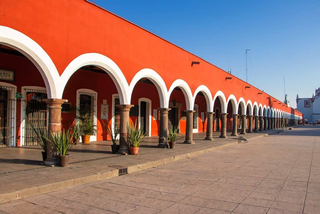 Colorful building with white arches and columns in a Mexican town, with potted plants and a clear blue sky.